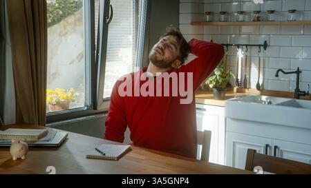 Müder junger Mann, der seinen Hals ausdehnt, während er an einem Küchentisch sitzt und sich nach langer Arbeit oder Studium erschöpft und müde fühlt Stockfoto