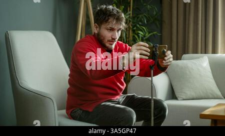 Mann in einem roten Pullover, der sein Smartphone auf einem Stativ aufstellt, während er in einem modernen Wohnzimmer sitzt Stockfoto