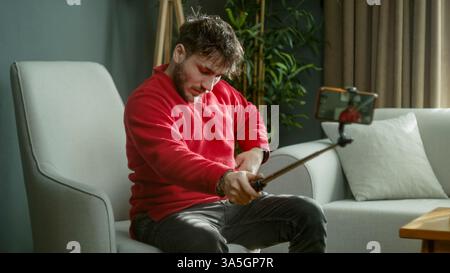 Mann in einem roten Pullover, der einen Selfie-Stick benutzt, um ein Video aufzunehmen oder ein Foto zu machen, während er in einem modernen Wohnzimmer sitzt Stockfoto