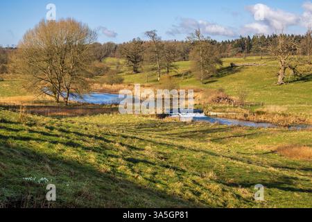 Fußgängerbrücke und Weir am Sherborne Brook in Sherborne Village, Cotswolds AONB, Gloucestershire, England Stockfoto