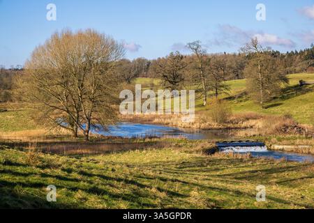 Fußgängerbrücke und Weir am Sherborne Brook in Sherborne Village, Cotswolds AONB, Gloucestershire, England Stockfoto