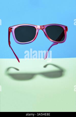 Sonnenbrille in leuchtend rosa Farbe in der Luft. In der Luft schwebende rosa Sonnenbrille auf pastellfarbenen blauen und grünen Hintergrund. Senken Sie den Schatten unter dem Glas Stockfoto
