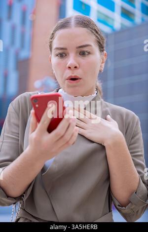 Schockierte kaukasische Geschäftsfrau, die draußen mit überraschtem Ausdruck auf das Smartphone blickt. Stockfoto