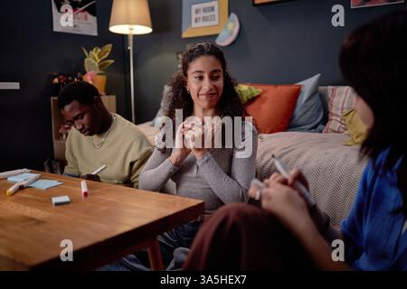 Lächelnde Frau, die auf der Couch sitzt und sich mit zwei Freunden um einen hölzernen Couchtisch in einem gemütlichen Wohnzimmer mit farbenfrohen Kissen und Postern unterhält Stockfoto