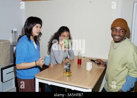 Drei Freunde genießen gemeinsam Drinks am Café-Tisch, unterhalten sich, einer hält das Telefon und lächelt. Zwanglose Atmosphäre mit Getränken und entspannter Atmosphäre Stockfoto