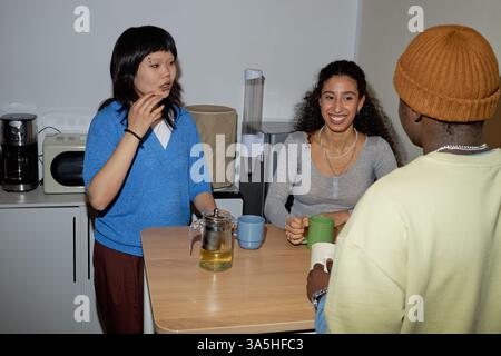 Eine Gruppe von drei Personen versammelte sich um den Küchentisch und genoss Tee und Gespräche. Hintergrund zeigt Küchengeräte und einfache Einrichtung Stockfoto