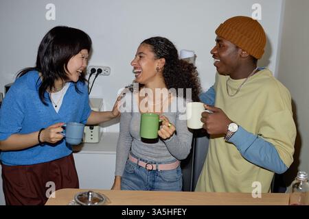 Eine Gruppe von Freunden, die in der Küche stehen und die Gesellschaft des anderen genießen und lachen, während sie Kaffeetassen halten, was eine warme und freundliche Atmosphäre schafft Stockfoto