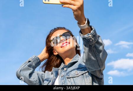 Junge Brünette, die eine Sonnenbrille trägt, macht Selfie mit ihrem Handy. Stockfoto