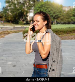 Junge Erwachsene städtische Frau lächelnd auf der Straße der Stadt zu laufen und am Sommertag mit dem Handy zu sprechen Stockfoto