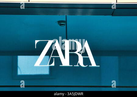 Eine Nahaufnahme des ZARA-Logos, weiß, auf einer Ladenfront aus blauem Glas mit Reflexionen und einer Sicherheitskamera, sichtbar in Manchester, Großbritannien. Stockfoto