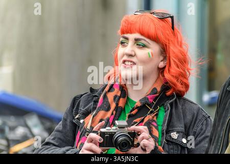 St. Patrick Parade. Arklow, Irland. 2025 Junge Frau mit leuchtend roten Haaren und irischem Make-up, die das festliche Ereignis mit einer Kamera festnimmt. Stockfoto
