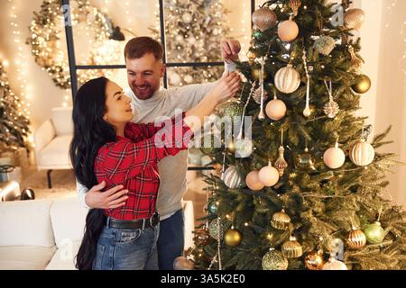 Sie schmücken den Weihnachtsbaum. Das Paar feiert zusammen das neue Jahr drinnen. Stockfoto