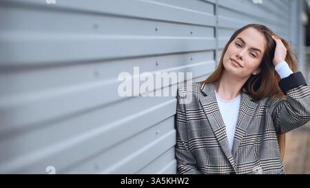 Junge Geschäftsfrau in karierter Jacke, die sich an eine graue Metallwand lehnt, ihr Haar berührt und lächelt Stockfoto