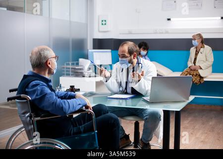 Ein junger Arzt im Laborkittel sitzt am Schreibtisch und spricht mit einem älteren Patienten im Rollstuhl mit Laptop und Klemmbrett in der Nähe. Ärztliche Beratung im Ruhestand kranker Mann, als Krankenschwester sitzt hinter einer assistierenden Seniorin. Stockfoto
