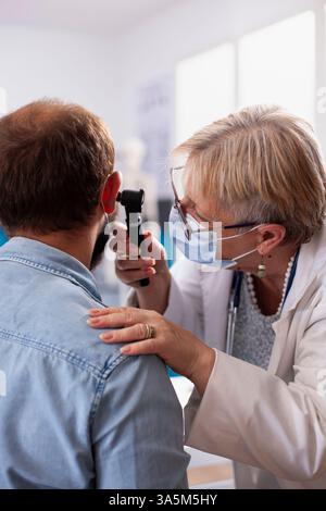 Der weiße Mann sitzt ruhig im Klinikraum, während ein hochrangiger hno-Spezialist sein Ohr auf mögliche Hörprobleme überprüft. Alte Ärztin mit Otoskop während der ärztlichen Untersuchung bärtiger männlicher Patienten. Stockfoto