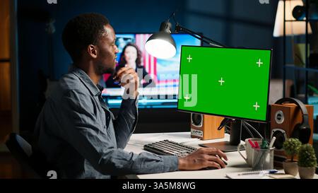 afroamerikaner, der von zu Hause aus arbeitet und den grünen Bildschirm am PC überprüft, hat einen Remote-Job in einem Heimbüro. Teleworker Multitasking und Networking in seinem modernen Loft. Kamera B.. Stockfoto
