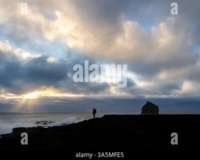 Eine Einzelfigur steht an der zerklüfteten Küste von Valahnúkamöl, Island, vor einem dramatischen Himmel mit Sonnenstrahlen, die durch stürmische Wolken brechen Stockfoto