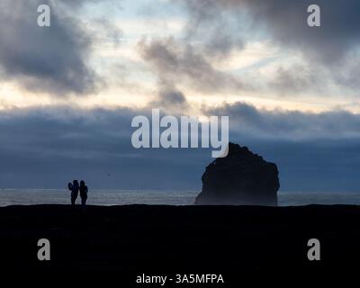 Zwei Reisende stehen auf einer Klippe mit Blick auf einen riesigen Meeresstapel vor Valahnúkamöl, Island, während Wellen brechen und Seevögel unter einem stürmischen Himmel schweben Stockfoto