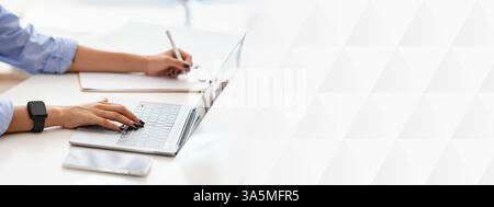 Professionelle Frau in blauem Hemd Multitasking, schreibt auf einem Laptop, während sie Notizen schreibt Stockfoto