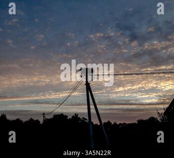 Storchennest auf einem Versorgungsmast bei Sonnenuntergang in einem ländlichen ukrainischen Dorf. Silhouette eines Vogelnest vor dramatischem Abendhimmel mit verstreuten Wolken. Stockfoto