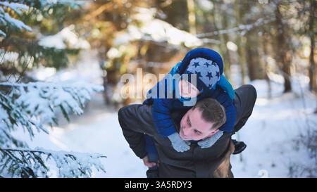 Vater trägt Sohn spielerisch auf dem Rücken, schneebedeckte Waldlandschaft mit majestätischen Immergrünen für eine friedliche Winterszene Stockfoto