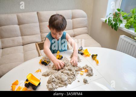 Kind, das zu Hause mit kinetischem Sand und Spielzeugbaufahrzeugen spielt. Aktivitäten im Innenbereich für kreatives Spiel und Entwicklung. Kind im Alter von drei Jahren Stockfoto