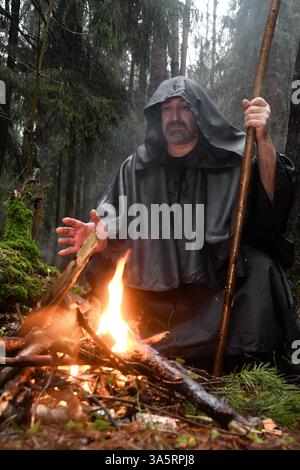 Ein Mönch in einem schwarzen Gewand sitzt im Wald am Feuer und wärmt seine Hände. Mystik und Magie, Zauberer und Hexen Stockfoto