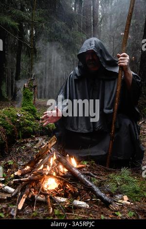 Ein Mönch in einem schwarzen Gewand sitzt im Wald am Feuer und wärmt seine Hände. Mystik und Magie, Zauberer und Hexen Stockfoto