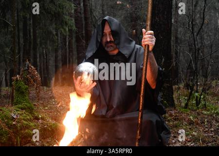 Ein Mönch in schwarzem Gewand sitzt am Feuer im Wald und schaut in einen Wahrsagungsball. Mystik und Magie, Zauberer und Hexen. Stockfoto