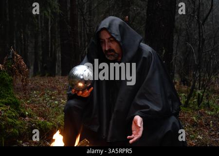 Ein Mönch in schwarzem Gewand sitzt am Feuer im Wald und schaut in einen Wahrsagungsball. Mystik und Magie, Zauberer und Hexen Stockfoto