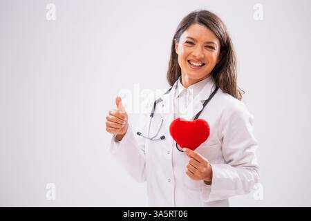 Das Studio hat ein Porträt einer fröhlichen Ärztin aufgenommen, die ein herzförmiges Kissen zeigt. Stockfoto