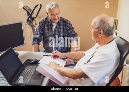 Glücklicher Senior-Mann macht eine Prüfung beim Kardiologen. Der Doktor sieht sich seine medizinischen Akten an. Glücklicher Arzt und Patient lächeln. Stockfoto