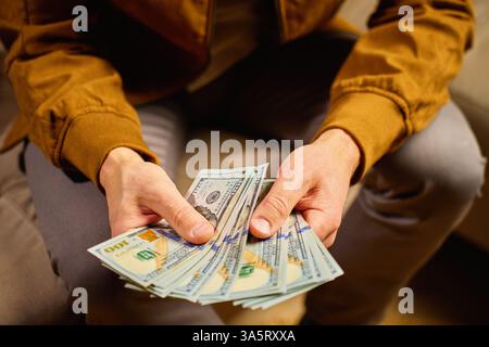 Person, die US-Dollar-Scheine zu Hause zählt. Mann, der auf dem Sofa sitzt und Bargeld in den Händen hält. Konzept von Geld, Sparen und Finanzplanung Stockfoto