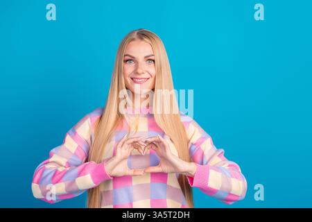 Porträt einer fröhlichen jungen Frau in einem farbenfrohen Pullover, der mit den Händen eine Herzgeste vor einem leuchtend blauen Hintergrund bildet Stockfoto