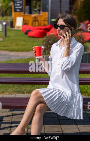 Stilvolle Frau in Sonnenbrille, die am Telefon spricht, während sie draußen eine Kaffeetasse hält Stockfoto