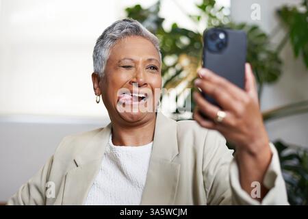 Fröhliche ältere Frau, die einen spielerischen Ausdruck macht, während sie für ein Smartphone Selfie im Haus posiert Stockfoto