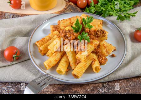 Teller mit Rigatoni-Pasta mit hausgemachter Bolognese-Ragu-Sauce auf Leinenserviette serviert auf rustikalem braunem Tischhintergrund Stockfoto
