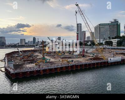 Luftaufnahme einer Baustelle in der Singapore Bay in der Dämmerung mit Baggern, Kränen und der Skyline der Stadt im Hintergrund. Stockfoto