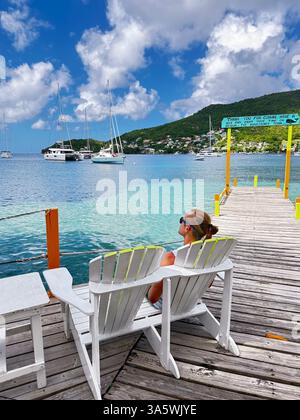Mann, der einen friedlichen tropischen Moment auf einem hölzernen Dock genießt, sitzt in einem weißen Stuhl mit Blick auf das türkisfarbene Meer, Segelboote und üppige grüne Hügel unter hellem Himmel. Stockfoto