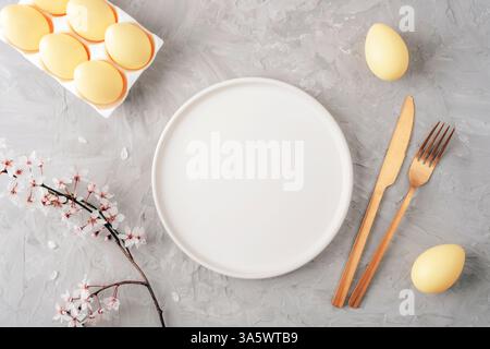 Leerer weißer runder Teller mit goldenem Besteck, gelben Eiern und Kirschblüten auf grauem Hintergrund. Osterkonzept. Draufsicht, flach, Modell. Stockfoto