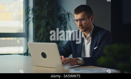 Konzentrierter Geschäftsmann im Anzug, der in einer modernen Büroumgebung an einem Laptop arbeitet. Ernsthafte, konzentrierte Ausdrücke deuten auf tiefgründiges Denken und Analyse hin Stockfoto