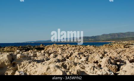 Wunderschöne Küstenlandschaft mit felsigem Gelände und klarem blauem Wasser unter einem sonnigen Himmel, mit grünen Hügeln und Bergen in der Ferne Stockfoto