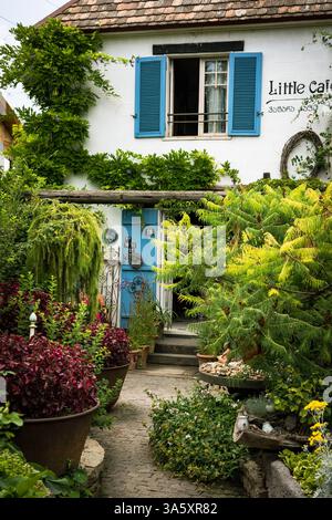 Ein bezauberndes altes Haus mit Terrakotta-Dach, blauen Fensterläden und weißen Putzwänden liegt eingebettet in einen üppigen Garten. Umgeben von Grün und Vibran Stockfoto