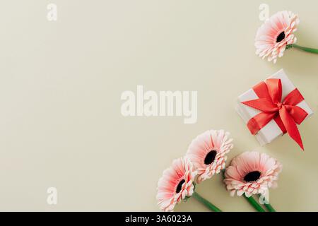 Geschenkbox mit rotem Band und rosa Gerbera-Blüten auf hellem Hintergrund. Draufsicht, flach, Kopierbereich. Stockfoto