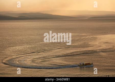 Ein Boot mit Passagieren auf ruhigem, goldenem Wasser bei Sonnenuntergang, ferne Inseln, trübe Sonne, Weitsicht. Kroatien. Stockfoto