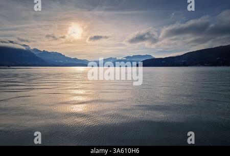 Ruhiger Gletschersee von Annecy mit sanften Wellen auf der Wasseroberfläche, die das sanfte Licht der untergehenden Sonne reflektieren. Die Aravis-Berge liegen im Stockfoto