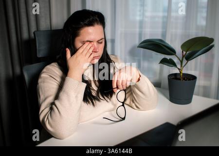 Müde Frau, die zu Hause mit einem Laptop arbeitet, der die Brille hält, die Augen reibt und Müdigkeit spürt. Stress- und Frustrationskonzept. Müde Augen. Stockfoto