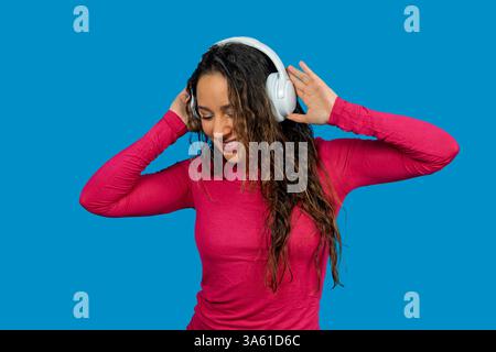 Glückliche junge Frau, die Musik mit kabellosen Kopfhörern hört, tanzt und lächelt auf blauem Hintergrund Stockfoto