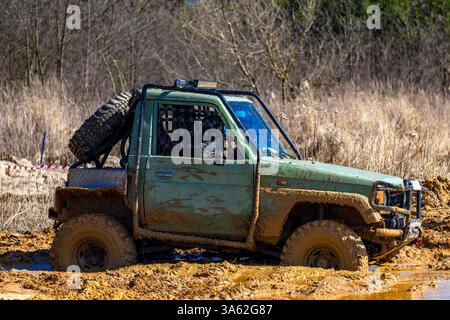 Ein grünes Geländefahrzeug steckt in einem riesigen Loch mit Schlamm und Wasser, überquert schwieriges Gelände mit dem Auto und tritt auf der Off-Road-Rennstrecke an Stockfoto