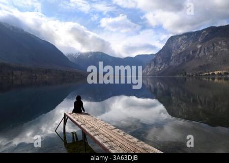 Frau, die im Winter auf einem hölzernen Dock mit Blick auf den See und die majestätische Berglandschaft mit sich spiegelnden Wolken im ruhigen Wasser sitzt, Bohinj-See Stockfoto